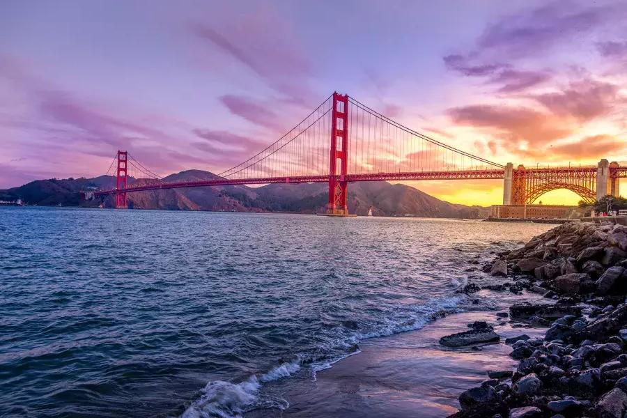 A Ponte Golden Gate ao pôr do sol com um céu multicolorido e a Baía de São Francisco em primeiro plano.
