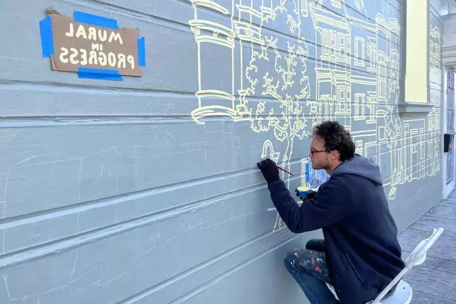 Um artista pinta um mural na lateral de um prédio no Mission District, com uma placa colada no prédio que diz "Mural em andamento". São Francisco, Califórnia.